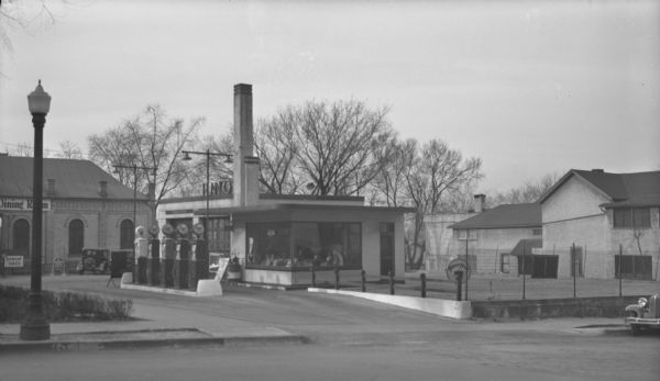 The Fiore Pennco gas station, 202 West Washington Ave at North Fairchild Street. There are five gas pumps, and on the left there are signs for Dunlop Tires and Quaker State Motor Oil in front of the Gates of Heaven Synagogue which has a sign for "Dining Room" along the side. To the right is a fenced-in empty lot, and in the background is a two-story building. At the entrance to the gas station is a sign that reads: "NevrNox," a brand of anti-knock compound.