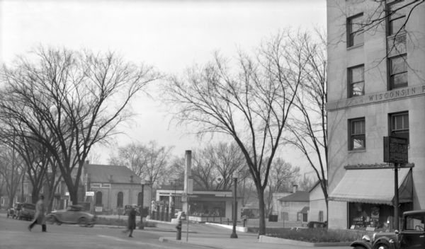 View across West Washington Avenue towards the Fiore Pennco gas station, 202 West Washington on the corner of North Fairchild Street. On corner on the right is a large stone building with the sign "Wisconsin P---," which is the Wisconsin Power and Light Company at 122 West Washington Avenue. In front of this building is a neon sign for the Union Bus Station, also at 122 W. Washington Avenue. In the background on the left beside the gas station is the Gates of Heaven Synagogue, with a sign on the side which reads: "Dining Room."