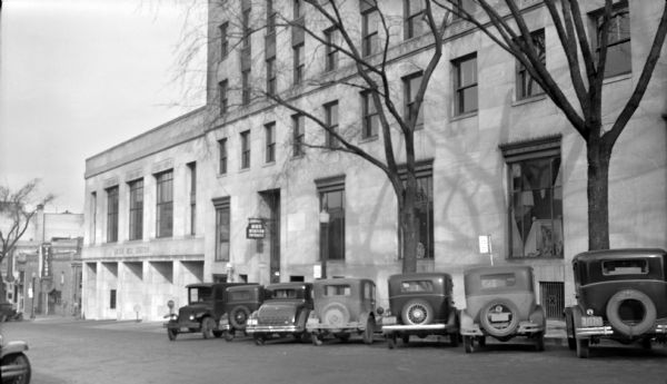 View across Fairchild Street towards the Union Bus Station at 122 W. Washington Avenue, with a row of automobiles parallel parked outside. On the exterior of the building is a barber pole for Mike's Barber Shop just below a sign that reads: "Bus Station Entrance." Down the street on the left is a "Tailor" sign on a building.