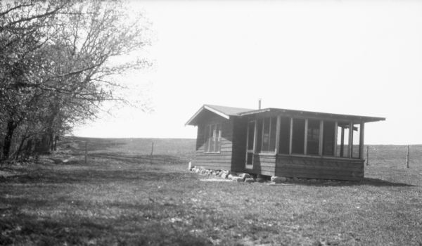 View up sloping lawn towards the Stanley Hanks cottage at Fox Bluff with a fence in the background. The cottage has a screen porch along one side and a rough stone foundation.