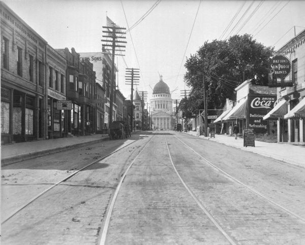 View up center of King Street towards the third Wisconsin State Capitol. There are streetcar tracks, and large power poles are along the left side of the street. A building on the corner of E. Main Street on the left has a "Smoke" sign. There is a horse-drawn vehicle and a few automobiles parked along the curbs. On the right is a store with a "Meals" sign next to a small grocery store with an awning advertising cigars and fruits. On the side of the building is a large painted advertisement for Coca-Cola. The building in the right foreground is a paint and wallpaper store with a large sign advertising "Patton's Sun Proof Paints," and awnings advertising glass and wallpaper. On the sidewalk is a signboard: "Emil O. Seiler Contractors and Painting: Interior wood finishing, calcimining, picture framing, decorating, glass glazing and varnishes, etc."