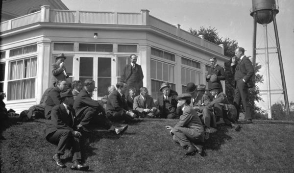 A large group of men in three-piece business suits standing and sitting together on the lawn at the Maple Bluff Country Club. There is a water tower in the background on the right.