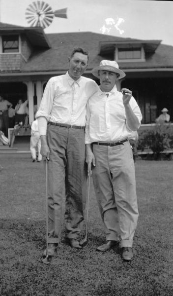 Portrait of two golfers wearing white shirts and smoking cigars posing outdoors. They clubs they are holding are drivers. Behind them people are relaxing on the porch of the country club. A water tower is in the background.