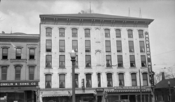 View from across street towards the upper facade of the Madison Hotel, on the corner of North Pinckney and Mifflin Streets. Storefronts in the building include Woldenberg's Women's Clothing, along with a cafeteria. Conklin and Sons Company, a coal business, is in the building next door on the left.