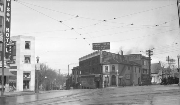 View from Mifflin Street towards the Hommel Brothers Restaurant at the corner of North Hamilton Street and East Mifflin Street. The restaurant started off as a grocery. A sign on the roof reads: "Hommel Bros Store, Groceries'. And a billboard on top: 'Hommel Bros., Good Things to Eat." On the far left are signs attached to the corner of the new Madison Hotel. There are trucks parked on Mifflin Street on the right.