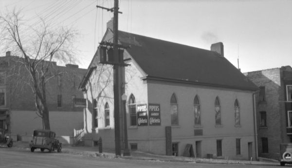 View across street towards Piper's Garden Cafeteria, at 120 East Mifflin Street, in the original building with church-style windows. There is a sign for a shoe repair shop leaning on the outside wall.