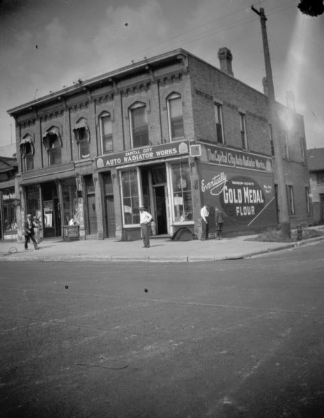 View from intersection at East Washington Avenue and Webster street, of the building at 121-123 E. Washington. The street has streetcar tracks. On the side of the building is painted advertisement: "Eventually Washburn-Crosby's Gold Medal flour. Why Not Now?." Pedestrians are on the sidewalk, and a young child is standing at the open door entrance to Capital City.