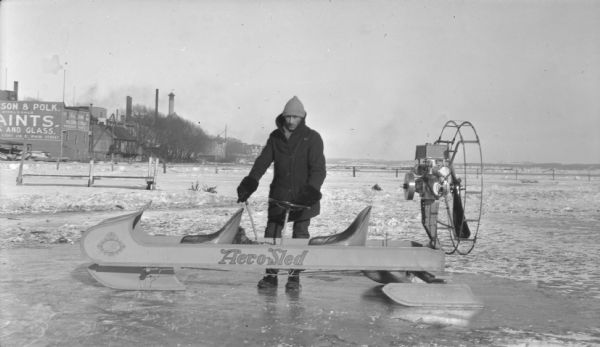 A man dressed in winter clothes is standing behind an Aero-Sled iceboat, which has a motor and a propeller on the back. The boat is near the Lake Monona shoreline. In the background is the Jackson and Polk Paints store at 218 East Main Street.