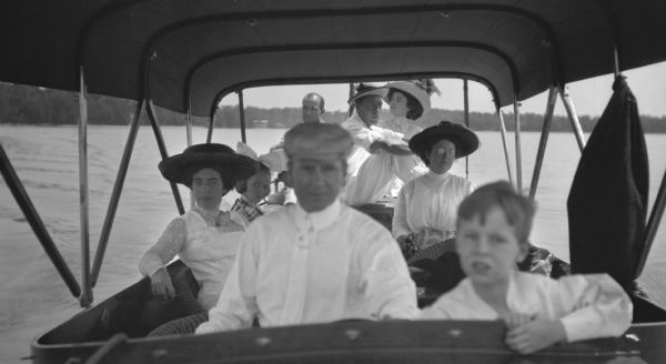 View from front of boat towards three men, three women, and a young girl and a boy riding in a launch. The boat cover shades the people in the front, while the three people sitting in the back are in the sun. The man piloting the boat, and the boy sitting next to him are out of focus.