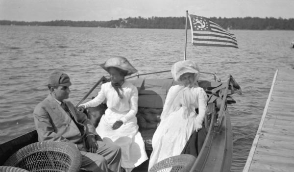 View from pier of a young couple sitting in a motorboat launch with an older woman. The two women are wearing hats with voluminous scarves and light-colored dresses. The man is wearing a suit and cap. Towards the front of the launch are empty wicker chairs. The launch has a flag, called a Yacht Ensign, with an anchor surrounded by a circle of stars. The far shoreline is in the background.