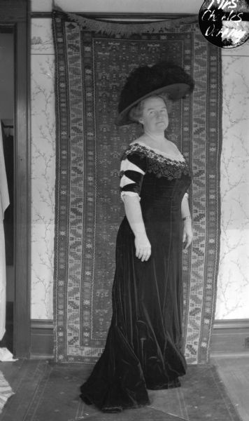 Full-length portrait of Mrs. Charles Derring standing and wearing a velvet dress and a large feathered hat. There is a carpet hung on the wall behind her as a backdrop.