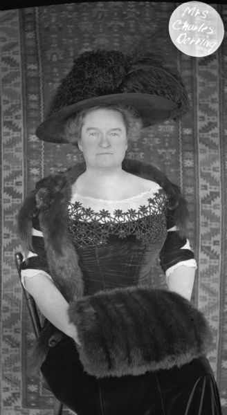 Three-quarter length portrait of Mrs. Charles Derring, sitting in a chair. She is wearing a velvet dress, feathered hat, fur stole and fur muff. Behind her a carpet has been hung on the wall as a backdrop.