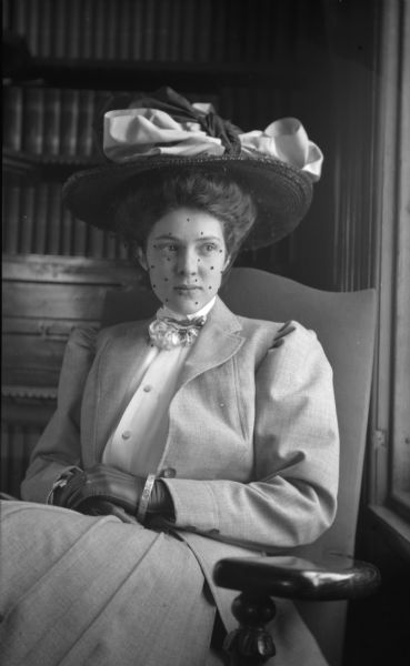 Sybil Nash, a Hanks family friend, is sitting in a chair near a window. She is wearing a beribboned hat with veil, a suit with a blouse and lacy tie, leather gloves and a bracelet.