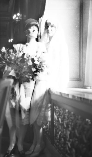 Full-length portrait of Sybil Nash, holding a bouquet of flowers and wearing a cloche hat, posing next to the bride. The bride is leaning on the window sill wearing a lacy veil.
