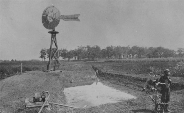 A man is standing in the right foreground near a tall plant, perhaps corn. In the center is a square-shaped pond with high banks. A man is in the background standing in a ditch that is diverting water into the pond. On the left is a pump with pipes going into and out of the pond, and behind the pond is a windmill.