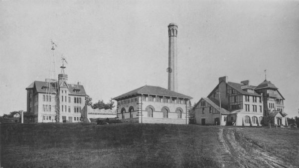 View across field towards Physics Hall, the heating plant, and Hiram Smith Hall. Physics Hall has two windmills; the heating plant has a large chimney, and the road to Hiram Smith Hall is not yet paved.