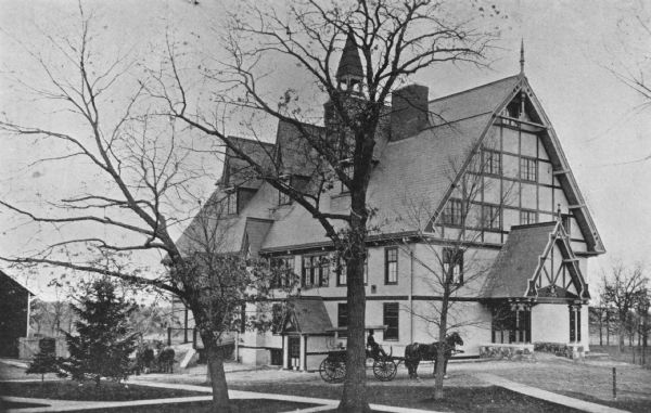 View of the horse barn. A man is driving a horse and buggy at the College of Agriculture on the University of Wisconsin campus. Two horses on standing on the left near the entrance of the barn. There is a central tower and half-timbering on the gable end of the upper stories of the barn. 