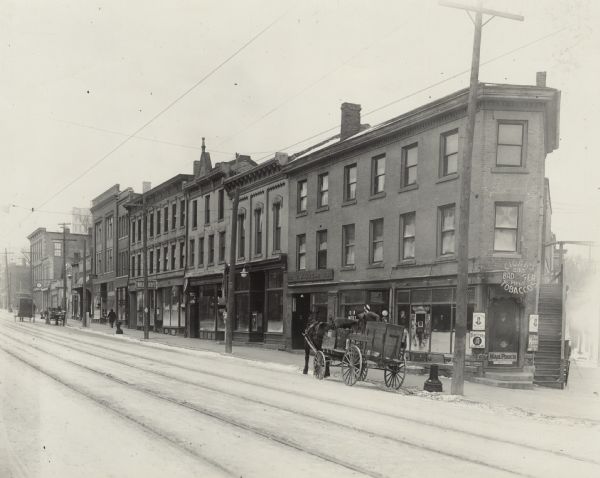 View across snowy street towards the 200 block of State Street, the north side. On the corner at number 235 is the Badger Cigar Pipes and Tobacco store. Moving to the left the next identifiable store is a barbershop with a striped pole and a painted sign on the window that reads: "U R Next," followed by C.J Kruse at number 231, selling fruit and bakery. Further down is a restaurant, grocery store, a sign for "S. Ribansky Ladies' Tailor," and a billiards hall. There are horse-drawn wagons or carts parked along the curb. 