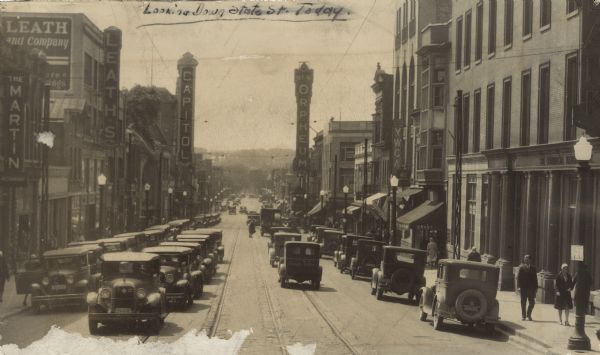 View of heavy automobile traffic, delivery trucks and pedestrians on State Street, seen looking west from the 200 block. In the background is Bascom Hill, on the University of Wisconsin-Madison campus. Commercial buildings include Leath's, Capitol Theatre, Orpheum Theatre, YWCA, the Commercial Bank, among others.