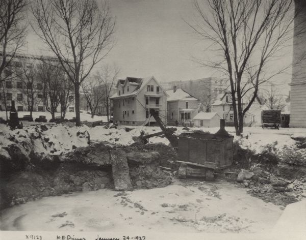 View over construction pit on the building site of the University of Wisconsin hospital. There is a steam shovel on the right far side. In the background are houses on North Charter Street, and beyond them Sterling Hall on the left and Chamberlin Hall on the right.