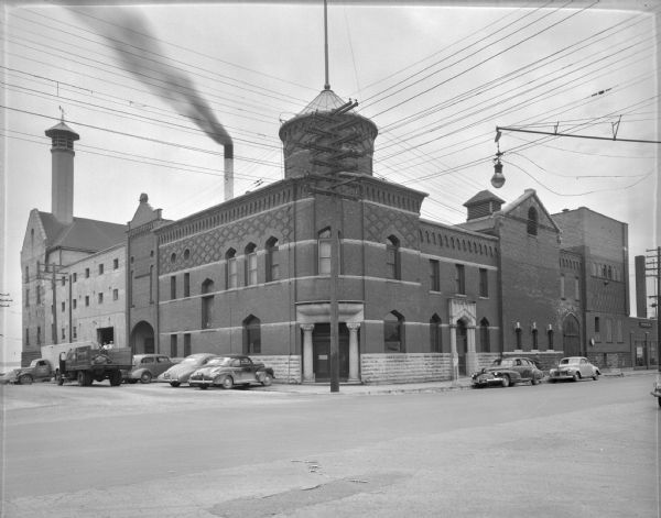 View across the street towards the Fauerbach Brewing Company, located at 651-653 Williamson Street. Automobiles are parked along the curb. On the left a truck is parked at the side of the building, and in the background is Lake Monona. There is a large electric power pole on the corner near the entrance to the brewery, and smoke is coming out of a smokestack behind the brewery.