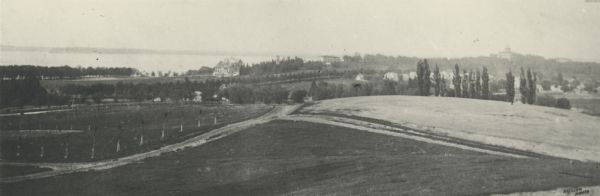 Slightly elevated view over fields of the University of Wisconsin-Madison, showing, from right, Bascom Hall, the Observatory, and King Hall (1525 Observatory Drive), from University Heights. Lake Mendota is in the background on the left. A neighborhood of homes are in the middle distance.