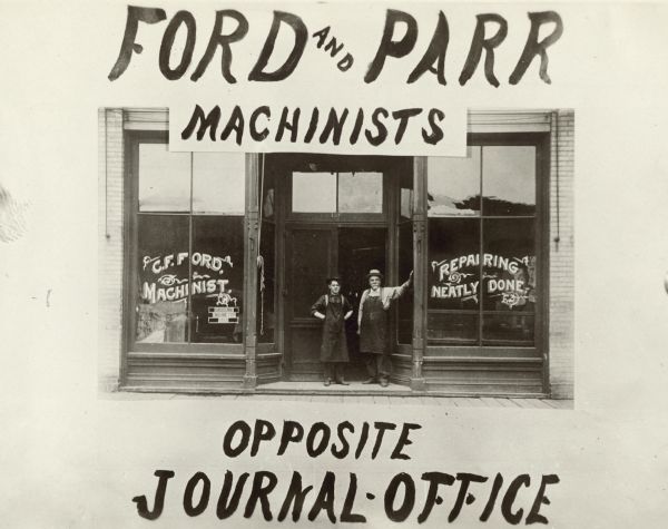 View from street of two men wearing work aprons posing in front of the Ford & Parr building at 120 East Washington Avenue. The painted sign on the window reads: "C.F. Ford, Machinist. Repairing Neatly Done." Written on the bottom: "Opposite Journal Office."
