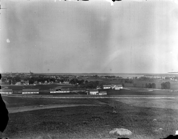 View from University Heights looking south across open fields towards barns and fences. (written on envelope: Camp Randall, Old Dane Country Fair Grounds). There is a long barn, or series of barns in the foreground. In the far background are houses, church buildings, and the Wisconsin State Capitol. On the far right is Monona Bay and Lake Monona.
