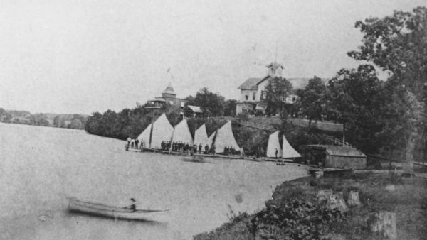 View along shoreline towards Tonyawatha House. A man or woman is in a rowboat in the foreground, and a large group of people are on a pier near five or six sailboats.