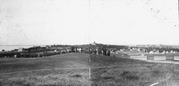 View from University Heights looking south across hills and open fields towards barns and fences. There is a series of long barns and a long fence on the right. Lake Mendota is on the far left, and Bascom Hill is on a hill in the center. In the far background on the right are houses, church buildings, and Lake Monona.