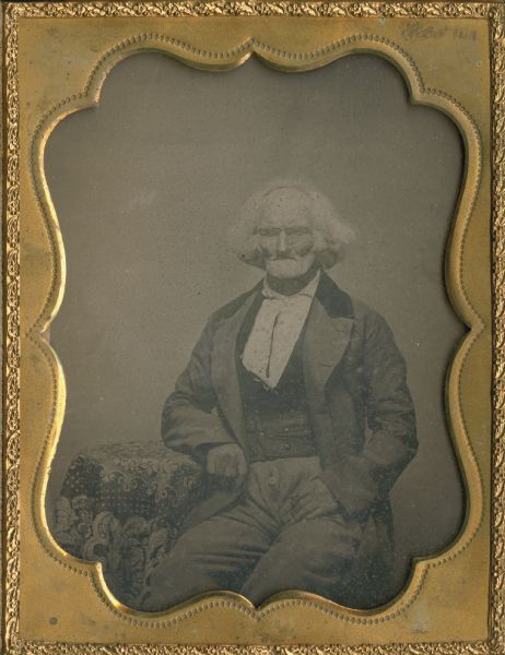 Quarter plate ambrotype. Three-quarter length studio portrait of James Steele sitting in a chair with his arm resting on a table covered with a patterned cloth.