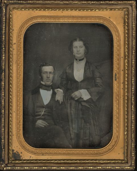 Half plate daguerreotype of an unidentified man and woman. The man is seated and the woman is standing and has her hand resting on the man's shoulder. The woman is wearing matching earrings, a necklace, and a brooch on a ribbon around her neck. Her dress has lace cuffs. The chain of the man's pocket watch is attached to his waistcoat. There is hand-coloring on the gold details of the woman's jewelry.