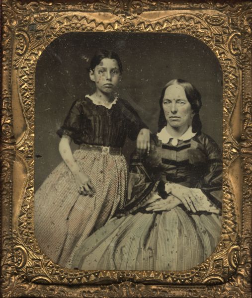 Sixth plate ambrotype portrait of a woman and a young girl. The woman is seated, and the girl is standing beside her resting her hand on the woman's shoulder. Hand-colored areas on face and dresses. This image was found among the effects of a dead Union Soldier during the Civil War and was donated to the collection by General Proudfit.