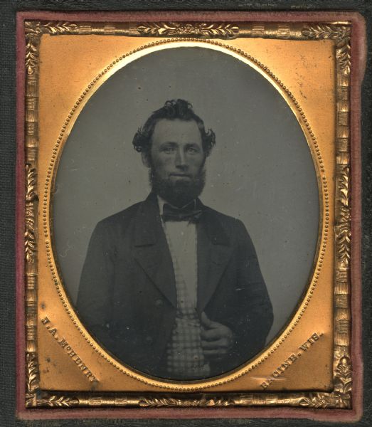 Sixth plate ferrotype/tintype of an unidentified man. Waist-up portrait facing forward, he is wearing a suit, bow tie, and checked vest. He is holding his the edge of his jacket with one hand. Hand-coloring on cheeks. 