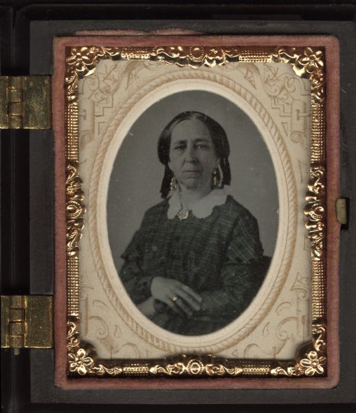 Ninth plate ferrotype/tintype of an unidentified woman. Waist-up portrait, she is facing forward with torso facing slightly left. She is wearing a plaid dress, cut-work white collar with brooch at the neck, drop earrings, a ring on her hand, and ribbons in her hair. Hand-coloring on gold jewelry details, and cheeks. 