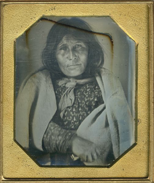 Sixth plate daguerreotype of an unidentified Native American woman, possibly from the Canadian Woodland Tribe. She is seated with a blanket around her shoulders and is holding an unidentified object in one of her hands.