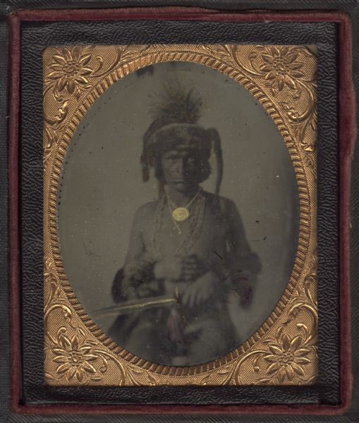 Sixth plate ferrotype/tintype of unidentified Native American man. Seated three-quarter length portrait, facing forward. He is wearing a fur and quill headdress and wristlets, a peace medal and beads around his neck, and is holding a feather-decorated unidentified object, possibly a pipe, in his hand. Hand-coloring of gold and red details on jewelry, headdress, wristlets, and pipe or dagger. 