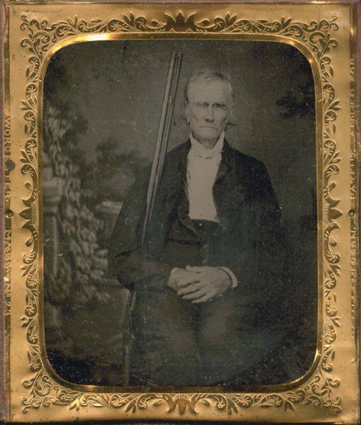 Sixth plate ferrotype/tintype. Three-quarter length portrait of Major John Gibson sitting in front of a painted backdrop, with his hands folded on his lap. He has his arm around a rifle which is leaning up against his shoulder. Hand coloring on cheeks.