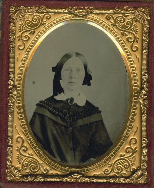 Ninth plate ambrotype. Waist-up portrait of Lucy Moody. Lucy is wearing a dark dress with a white-work collar, and a brooch at the neck. Over the dress she wears a wrap with ruffled trim and flowing sleeves. Her hair is parted down the center and large ribbons hold her hair up on both sides of her face. Hand coloring on cheeks. Lucy was the sister of J.L. Edwards. 