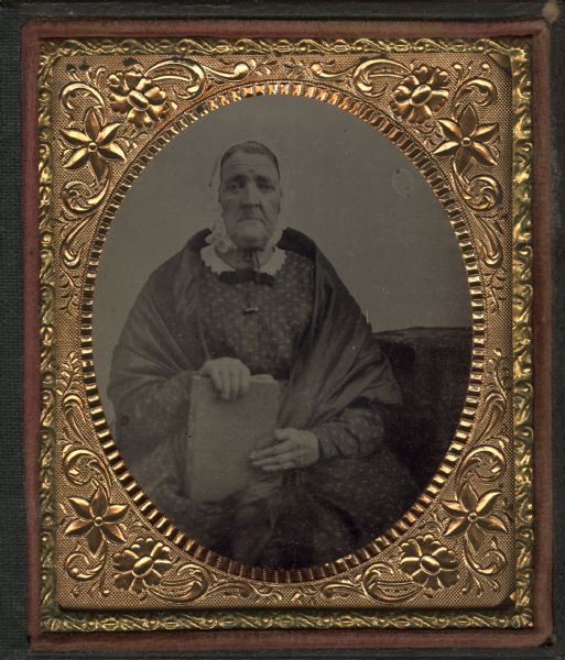 Sixth plate ferrotype/tintype of woman sitting and holding a book upright in her lap next to a cloth covered table. She is wearing a shawl over her dress and wears a white cap tied under her chin.