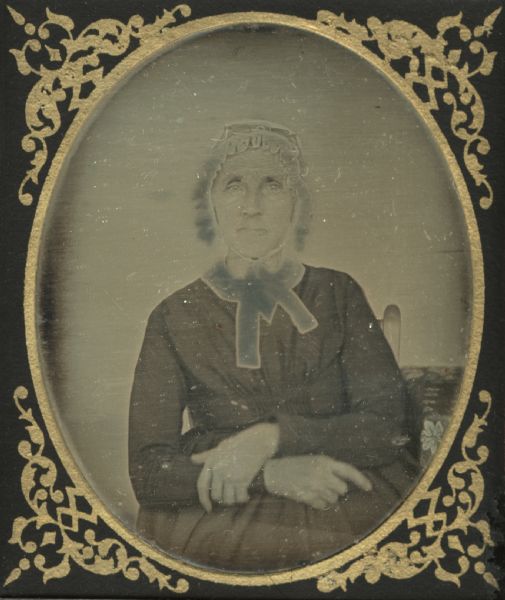 Sixth plate daguerreotype portrait of a woman identified as Aunt Churchill. The woman is seated in a chair with her arms folded in her lap. She is wearing a lace cap, and a pair of eyeglasses are resting on the top of her head. Hand-coloring on cheeks.