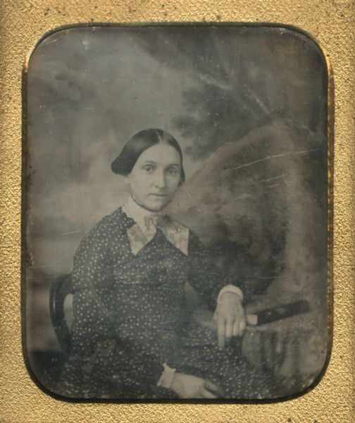 Sixth plate daguerreotype waist-up portrait in front of a painted backdrop of Mrs. Christian Miller. Mrs. Miller is seated in a chair with one arm resting on a book on a table.
