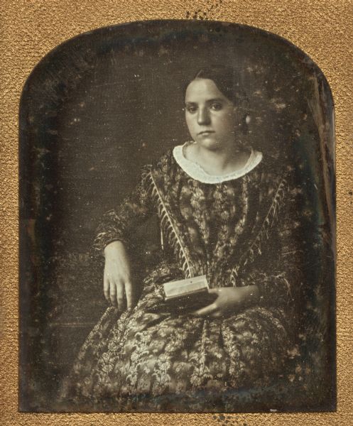 Sixth plate daguerreotype portrait of Celestia Mills. Miss Mills is seated with one arm resting on a table, and holding an open book with her other hand. Miss Mills was from Ohio and the niece of Simeon Mills, an early settler of Madison. Hand-coloring on the cheeks.