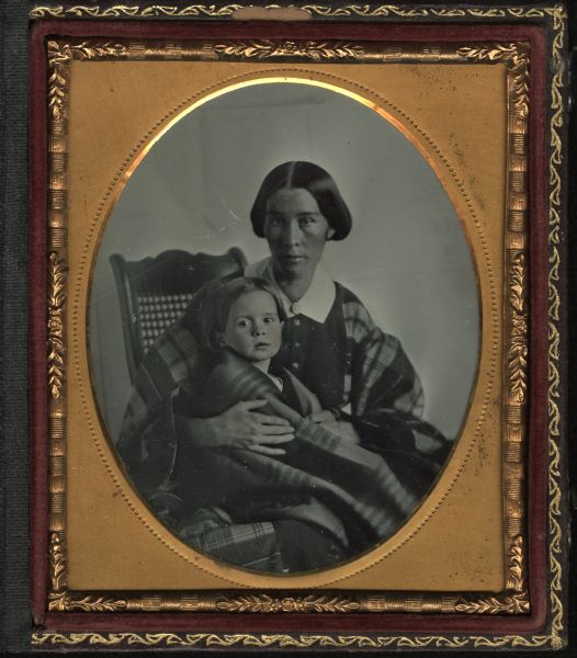 Sixth plate ambrotype portrait of unidentified woman seated on a cane-back chair, holding a child on her lap. The woman has a plaid shawl wrapped around her shoulders, and the child is wrapped in a striped shawl or blanket. Hand-coloring on cheeks.