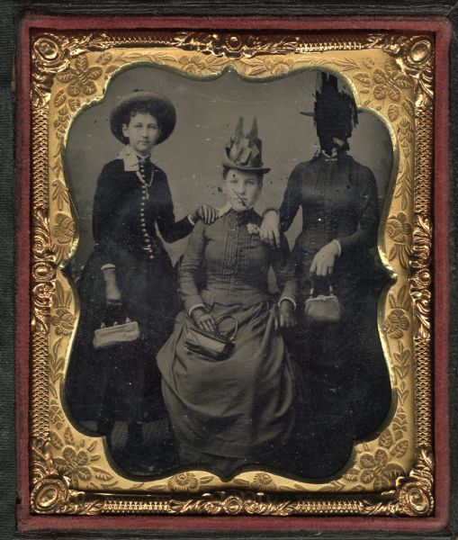 Sixth plate ferrotype/tintype of a full-length portrait of three women posing together. The woman in the center is sitting, flanked by two women standing, who each are resting a hand on the shoulder of the seated woman. The women are wearing dark jackets, skirts and hats, and holding small handbags. The women on the left and in the center are wearing gloves. The face of the woman on the right is deliberately scratched out. 