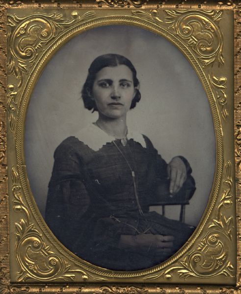 Sixth plate ferrotype/tintype of Mary Oaks. She is seated in a chair with one arm draped over the back and the other hand in her lap. She wears a long necklace that is attached at the bottom at her waist, perhaps for a watch, rings on her fingers, and a collar pin. Hand-coloring on cheeks and lips.