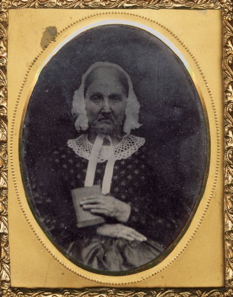 Sixteenth plate ferrotype/tintype of an old woman. She is seated with her hands in her lap and is holding a book. She is wearing a white bonnet tied with a ribbon under her chin. The woman has been possibly identified as the mother of Colonel P. Ege. Hand-coloring on cheeks.