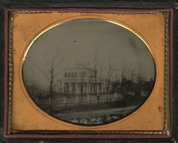 Quarter plate ambrotype exterior view of the Charles Minton Baker residence in Lake Geneva. The home was built in 1854. The house and grounds are shown from across the nearest road. Two women are standing on the front porch of the home and a man is standing near the fence of the property. To the right is a single horse and carriage.