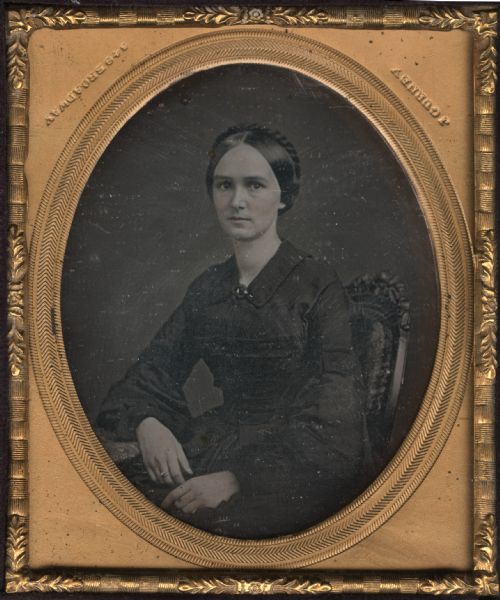 Sixth plate daguerreotype of Mary Louise Baker Lidgerwood Browne. Daughter of Charles Minton Baker of Lake Geneva. Taken in New York before her family journeyed to Wisconsin. She is in mourning dress, seated in an ornate wooden chair with her elbow resting on a table. Her hairstyle nearly covers her ears and is close fitted with a coronet of braid. Hand-coloring on cheeks and lips.