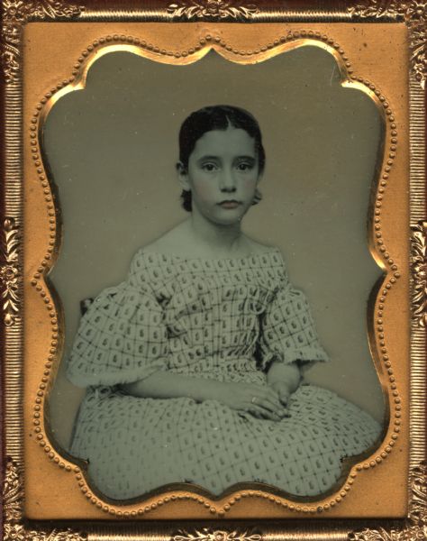 Ninth plate ambrotype of a young girl, most likely of the Charles Minton Baker family. She is sitting in a chair with her hands in her lap. Hand-coloring on cheeks and lips, and gold details on the ring on her right hand.